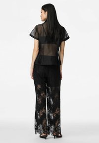 Kobieta ubrana w przezroczystą czarną bluzkę z krótkim rękawem i szerokie czarne koronkowe spodnie, stojąca tyłem do kamery na białym tle.