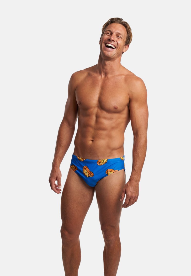 Hombre musculoso sonriente que lleva un bañador azul con un patrón de tigre naranja, de pie contra un fondo blanco liso.