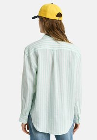 Camisa a rayas verde claro y blanca con mangas largas, puños abotonados y dobladillo curvo. Combinada con una gorra amarilla y azul marino.