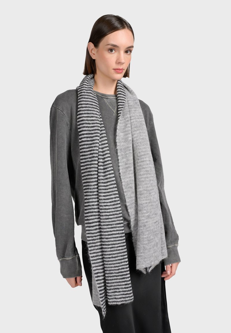 Femme aux cheveux bruns raides portant un haut à manches longues gris et une écharpe bicolore, un côté à rayures noires et grises, l'autre côté d'un gris clair uni.