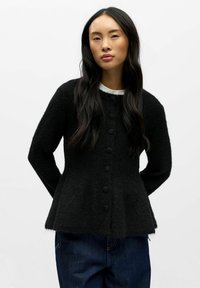 Musta neulottu neuletakki, jossa on teksturoitu pinta, istuva malli, pyöreä pääntie ja nappikiinnitys edessä. Päällä valkoinen paita.