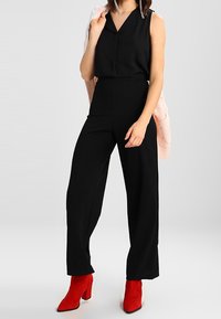 Haut noir sans manches avec un col, pantalon noir à jambes larges taille haute, et bottines rouges. Pull en maille rose pâle posé dessus.