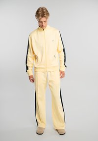 Joven con chándal amarillo con franjas negras a los lados, de pie y mirando hacia abajo sobre un fondo gris liso.