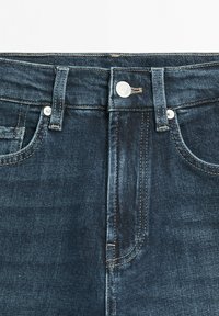 Dunkelblaue Jeans aus Denim im Nahaufnahme, die den Bund, die Gürtelschlaufen, den vorderen Knopf, den Reißverschlussbereich sowie die Fronttaschen mit Nieten auf weißem Hintergrund zeigen.