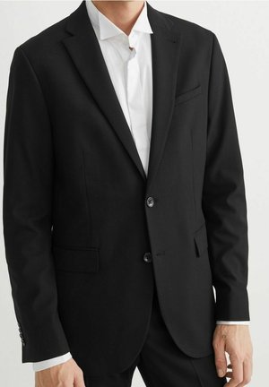 Mies, joka pukeutuneena räätälöityyn mustaan pukutakkiin ja valkoiseen kauluspaidan, seisoo yksinkertaista vaaleaa taustaa vasten.