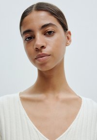 Close-up of a person with smooth skin and a neutral expression, wearing a textured, light cream top with a V-neckline and ribbed pattern.