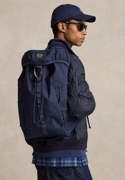 Άνδρας με σκουρόχρωμα γυαλιά ηλίου, ναυτικό καπέλο, μπουφάν με ραβδώσεις και καρό πουκάμισο, κυματίζει μια μεγάλη ναυτική τσάντα πλάτης σε απλή μπεζ φόντο.