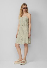 Blumengemustertes, hellgrünes Kleid mit tailliertem Schnitt, Spaghettiträgern und durchgehendem Knopfverschluss. Getragen mit klobigen beigen Sandalen.