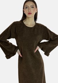 Vestido de pana marrón con escote redondo y mangas oversized. Presenta una textura suave y un ligero volante en los puños de las mangas.