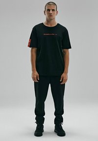 Czarna bawełniana koszulka z krótkim rękawem, czerwona tekst na przodzie i naszywka metkowa na prawym rękawie; połączona z czarnymi spodniami i trampkami.