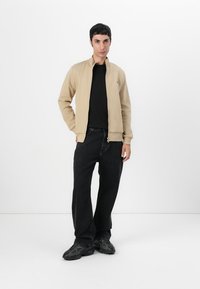 Beige zip-up jakke med ståkrage, parret med en sort t-shirt og sorte vide jeans. Tekstureret stof uden synlige mønstre.