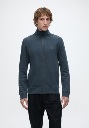 Joven con pelo rizado que lleva una chaqueta azul oscuro con cremallera y pantalones negros, de pie frente a un fondo liso y claro.