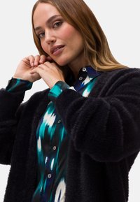 Svart fluffig cardigan över en skjorta med mönster i blått och turkost med knappdetaljer. Närabild av händer nära ansiktet. Släta texturer och färger.