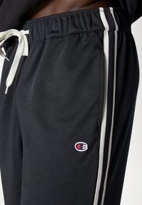 Musta collegehousut, joissa on teksturoitu pinta, joustava vyötärönauha, valkoinen nyöri ja kontrastiväri valkoiset pystysuorat raidat. Sisältää logopatchin.