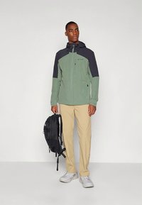 Zelena in črna softshell jakna s kapuco, žepi z zadrgo in lahkim materialom. Model nosi bež hlače in drži črno nahrbtnik.