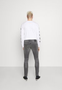 Camisa branca de manga longa com acentos de logotipo preto nas mangas, combinada com jeans cinza ajustados com painéis texturizados nos joelhos e sapatos pretos.