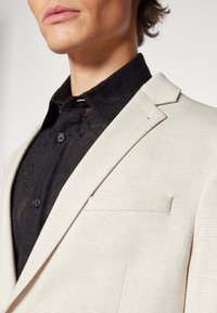 Beige rutet blazer med skarpe lapeller og en belagt lomme, kombinert med en svart teksturert skjorte med knappet krage og intrikate mønstre.