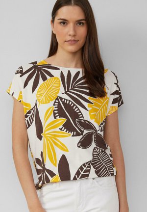 Jeune femme portant une chemise blanche avec de grands motifs de feuilles tropicales jaunes et marron et un pantalon blanc, se tenant devant un fond gris uni.