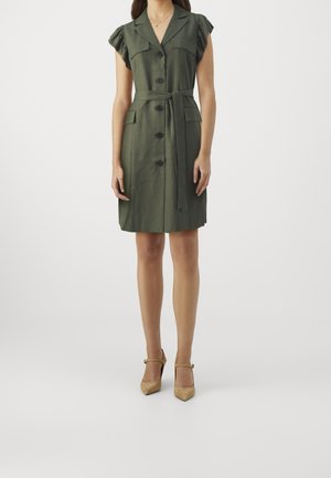Femme portant une robe ceinturée vert olive à manches courtes avec des boutons sur le devant et des talons à bride beige, debout devant un fond uni.
