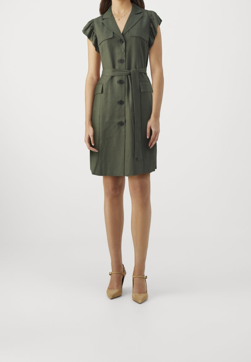 Femme portant une robe ceinturée vert olive à manches courtes avec des boutons sur le devant et des talons à bride beige, debout devant un fond uni.