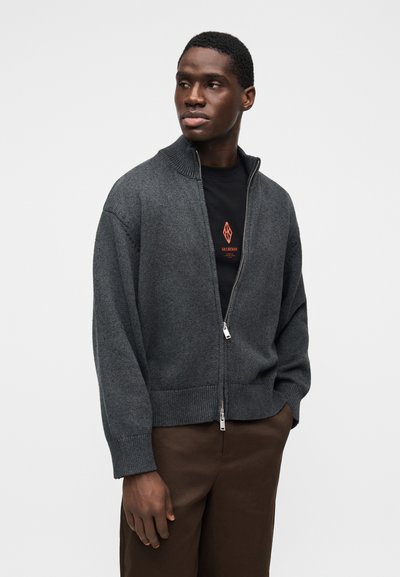 Muž v tmavosivej mikine na zips, cez ktorú má čierne tričko s potlačou, a v hnedej nohaviciach, stojaci pred jednoduchým svetlým pozadím.