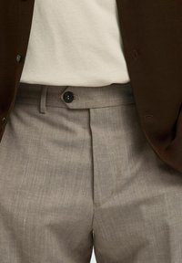 Detaljbild av en person som bär ljusbruna skräddarsydda byxor med en knapp och bälteshällor, i kombination med en vit skjorta och en brun jacka.