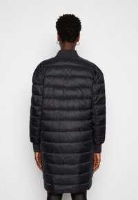 Abrigo de plumón largo y negro con un diseño acolchado, puños acanalados y cuello alzado, con una textura exterior lisa y brillante.