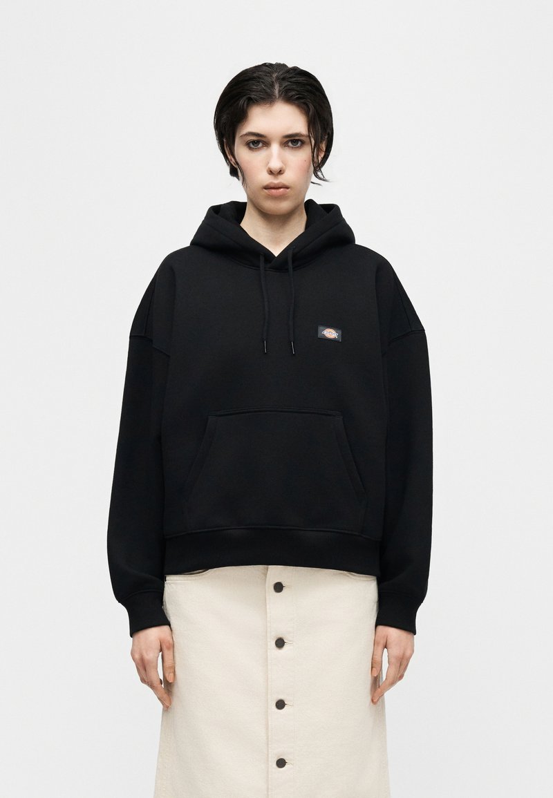 Person wearing a black hoodie with a front pocket and logo, paired with a beige buttoned skirt, standing against a plain light background.