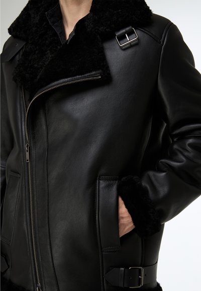 Schwarze Lederfliegerjacke mit Fellkragen, Reißverschluss und Schnallenakzenten. Texturierte Oberfläche mit weichem Fellbesatz an den Bündchen.