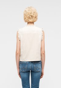Personne aux cheveux blonds courts et bouclés, portant un haut blanc sans manches à volants et un jean bleu, debout, dos au spectateur.