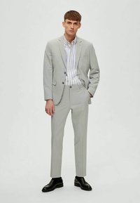 Homem em pé, vestindo um fato cinza claro, uma camisa de botão riscada e sapatos pretos contra um fundo claro simples.