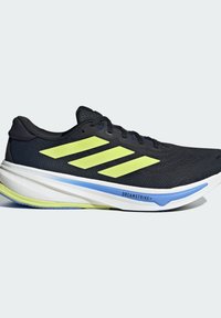 Zapatilla deportiva negra con tres franjas amarillas neón, punta redondeada, parte superior de malla texturizada y una media suela blanca con un acento azul.