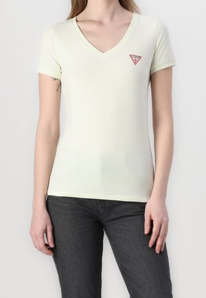 Kobieta ubrana w dopasowaną jasnżółtą koszulkę z krótkim rękawem i dekoltem w serek, z czerwonym logo Guess na piersi, w połączeniu z ciemnoszarymi dżinsami.