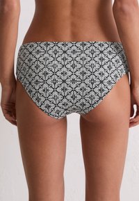 Mujer vistiendo una parte inferior de bikini con un estampado floral en blanco y negro, vista desde atrás sobre un fondo claro y liso.