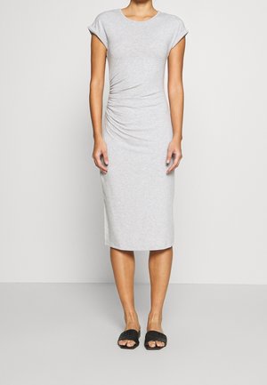 Kvinde iført en lysegrå, kortærmet, rynket midi-kjole med sorte åbne flade sandaler, stående mod en ensfarvet baggrund.