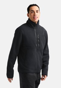Giacca in felpa a coste nera con colletto alto, tasche con zip e vestibilità affusolata. Presenta una superficie texturizzata per calore e comfort.