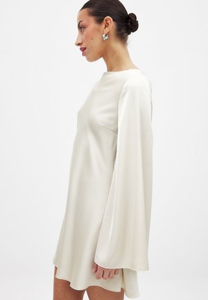 Vrouw met donker haar in een knot, draagt zilveren oorringen in de vorm van een cirkel en een loszittende, lange, crèmekleurige satijnen jurk, staande tegen een effen lichte achtergrond.