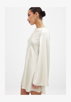 Sieviete ar tumšiem saknētiem matiem sasietiem bulciņā, valkājot sudrabotiem stīpas auskariem un brīvi pieguļošu, garām piedurknēm, krēmkrāsas satīna kleitu, uz vienveidīgas gaišas fona.