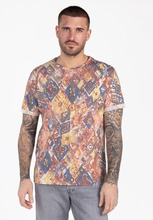 Homme aux cheveux courts et tatouages sur les deux bras, portant une chemise à motifs multicolores et un jean gris, debout devant un fond blanc.