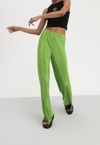 Pantaloni a gamba larga plissettati verdi con superficie testurizzata, abbinati a un top senza maniche nero con una grafica circolare. Sandali con piattaforma neri.
