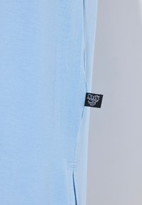 T-shirt azzurro chiaro realizzato in tessuto morbido, con una fessura laterale e un'etichetta nera cucita nella cucitura. Texture liscia e prominente.
