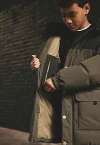 Giacca puffer con un esterno verde oliva e dettagli neri, dotata di una tasca interna con zip, bottoni a pressione e polsini elasticizzati.