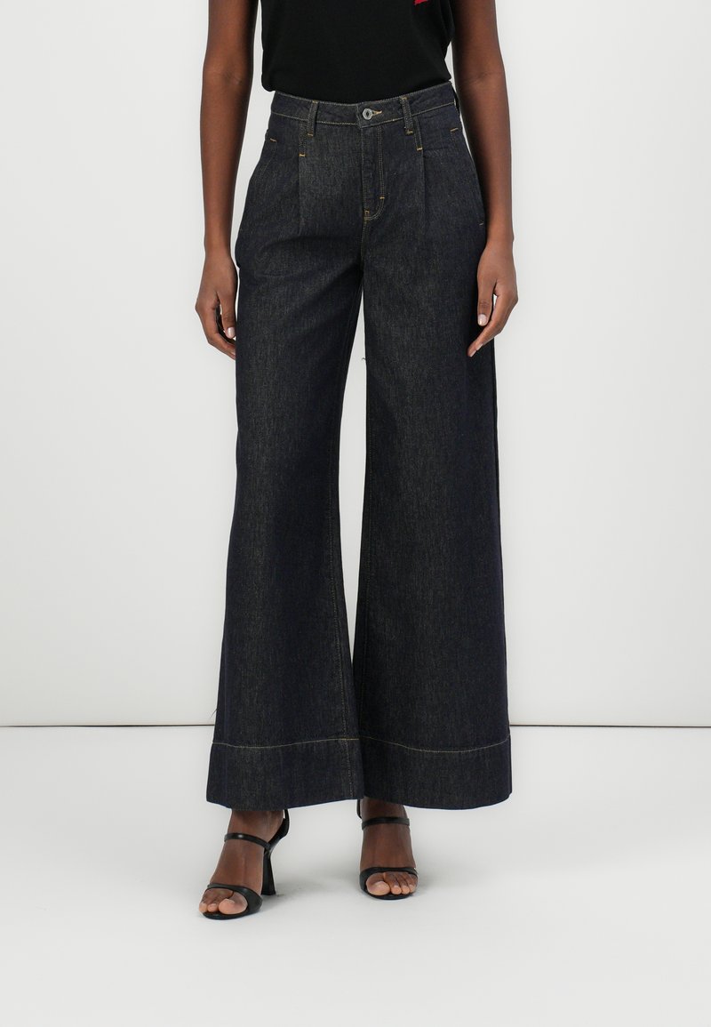 Dunkelblaue weite Jeans mit hoher Taille, ausgestattet mit zwei Fronttaschen und kontrastierendem gelben Nähten. Der Stoff hat eine glatte Textur.