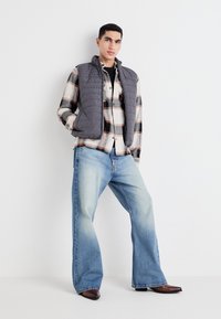 Chaleco acolchado gris sobre una camisa de cuadros con botones en rojo y negro, combinado con jeans de pierna ancha de color azul claro y botas marrones.