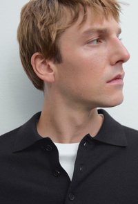 Young man with light brown hair wearing a black buttoned shirt over a white t-shirt, looking to the right against a plain background.