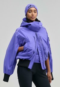 Lila vízálló dzseki túlságosan nagy kapucnival, több cipzáras zsebbel, elasztikus mandzsettákkal és fekete derékpánttal; fekete nadrág fölött viselve.