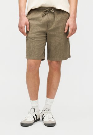 Άνδρας με υφασμάτινη χακί βερμούδα με κορδόνι στη μέση, λευκές κάλτσες μέχρι τον αστράγαλο και λευκά αθλητικά παπούτσια με μαύρες ρίγες, στέκεται μπροστά σε απλό φόντο.