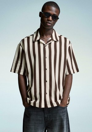 Short-sleeve button-up shirt with horizontal brown and cream stripes; textured fabric; relaxed fit; worn with dark jeans and round sunglasses.
