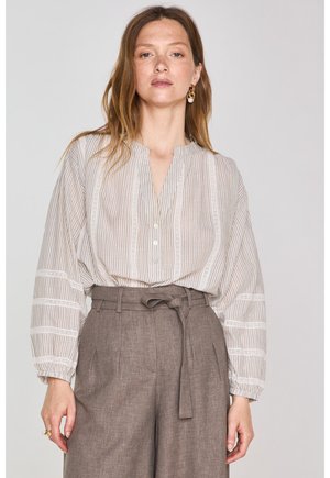 Blouse légère à rayures beige avec des garnitures en dentelle blanche, présentant un col en V et une patte de boutonnage, associée à un pantalon marron taille haute.