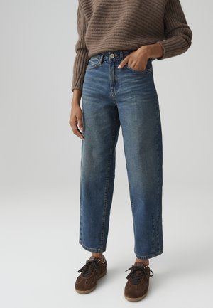 Persoon die hoge blauwe rechte jeans draagt, een bruine gebreide trui en bruine veter-sneakers, staand met één hand in de zak.